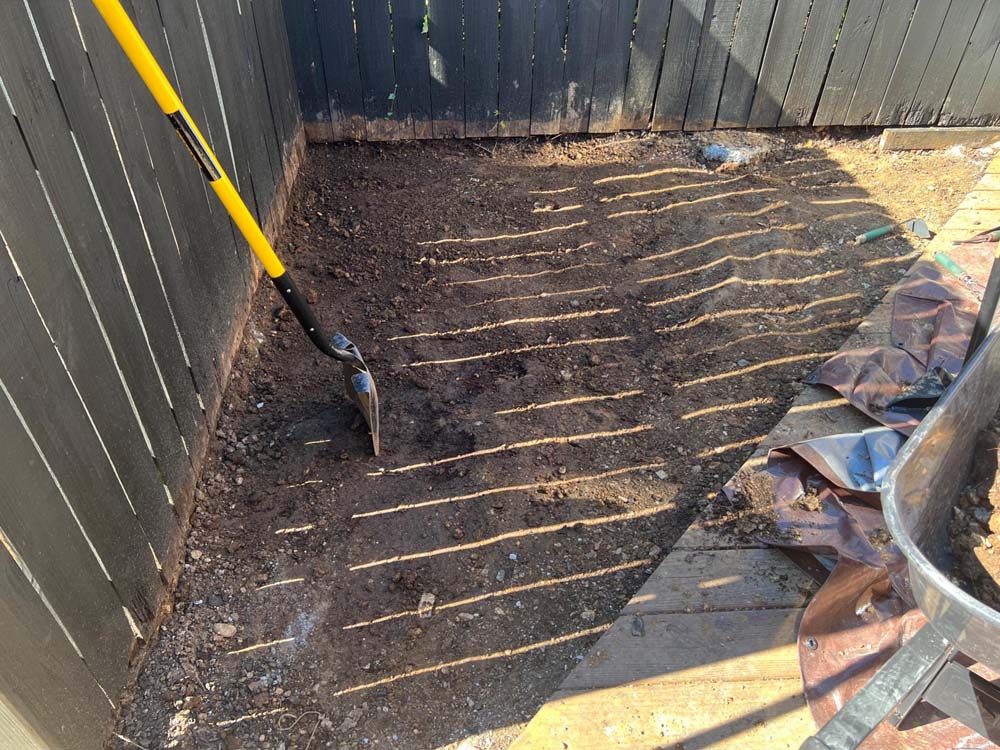 Corner of outdoor patio with dirt exposed, yellow shovel leaning up against dark fence.