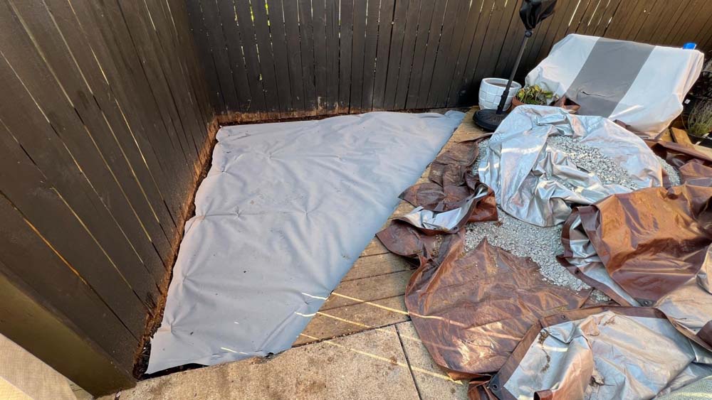 Corner of outdoor patio covered with grey tarp; small grey gravel on brown tarp sitting to the right of tarped corner. 