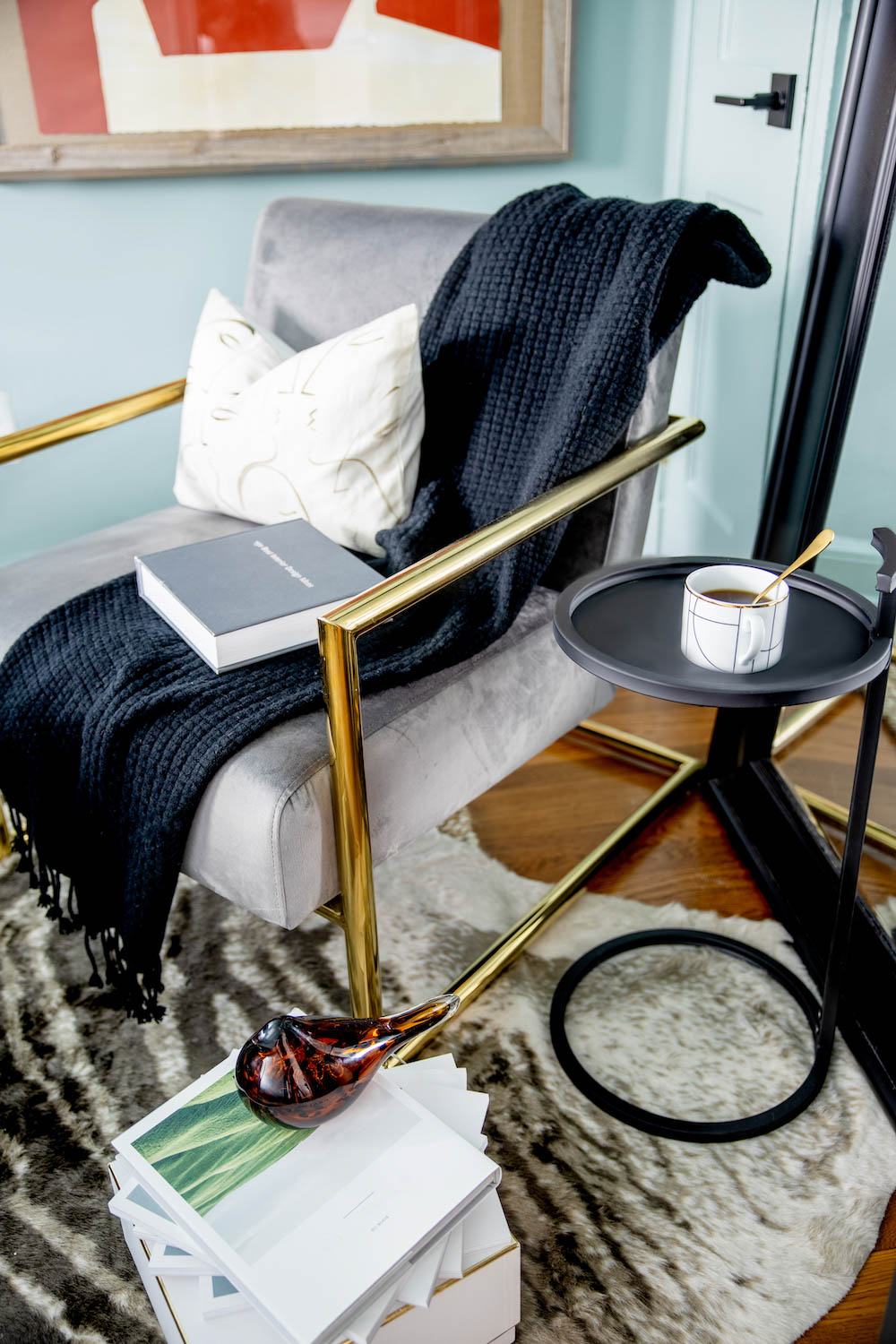 A gray velvet accent chair with gold accents next to a small black side table. A gray velvet accent chair with gold accents next to a small black side table.