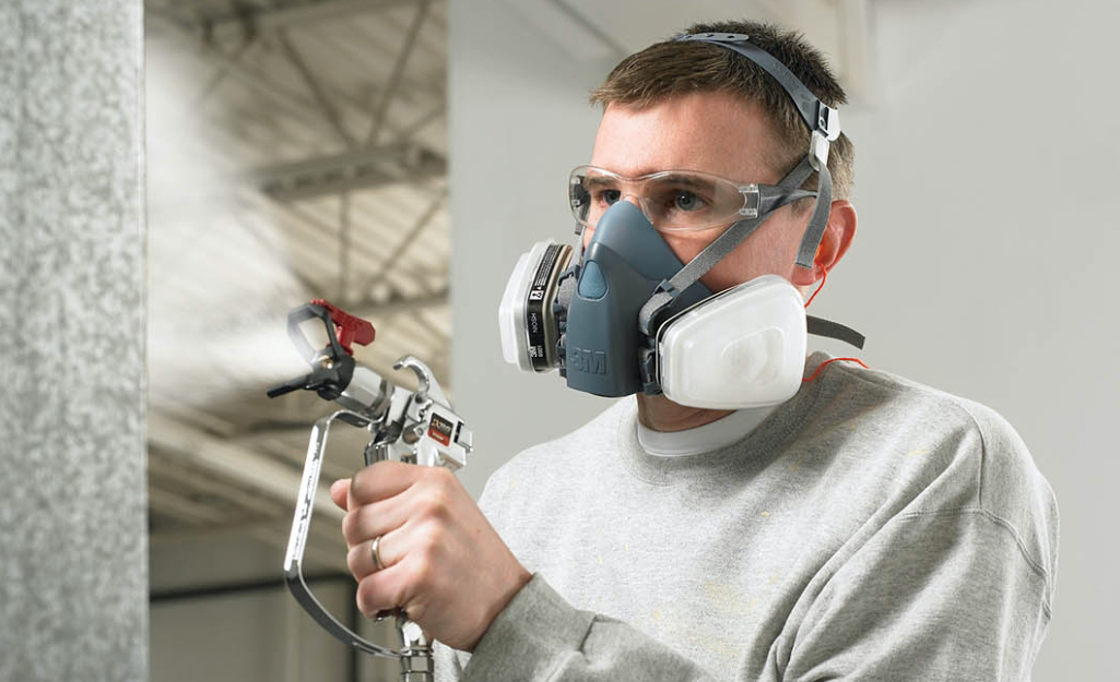 A man uses a paint sprayer while wearing a respirator.