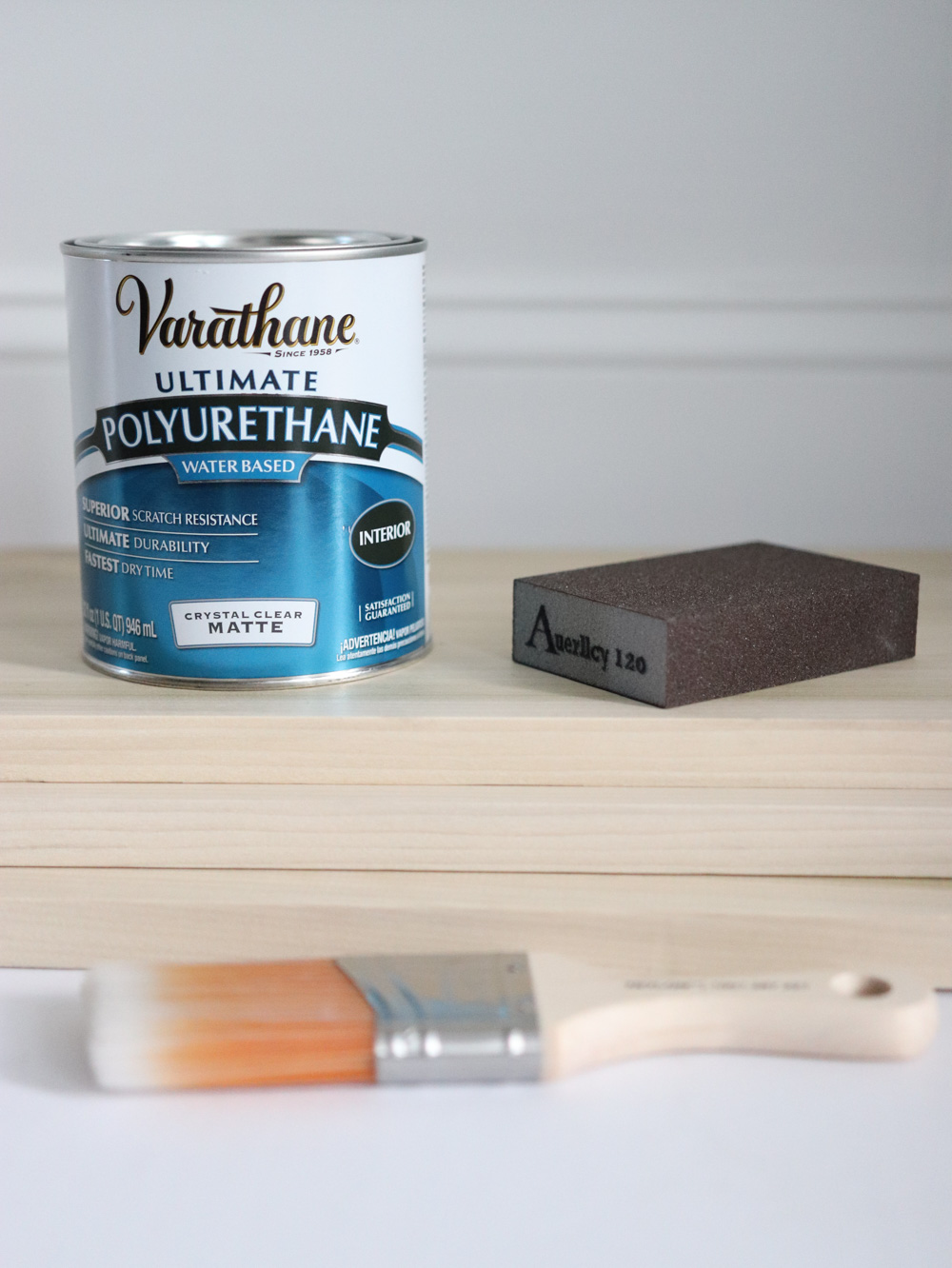 A can of Varathane polyurethane, a paintbrush, and a sanding block. A can of Varathane polyurethane, a paintbrush, and a sanding block.