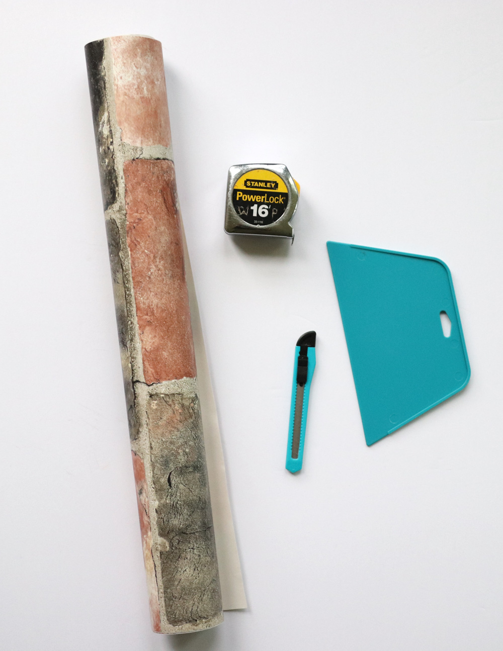 A roll of brick peel-and-stick wallpaper laid next to a wallpaper smoother and utility knife kit. A roll of brick peel-and-stick wallpaper laid next to a wallpaper smoother and utility knife kit.