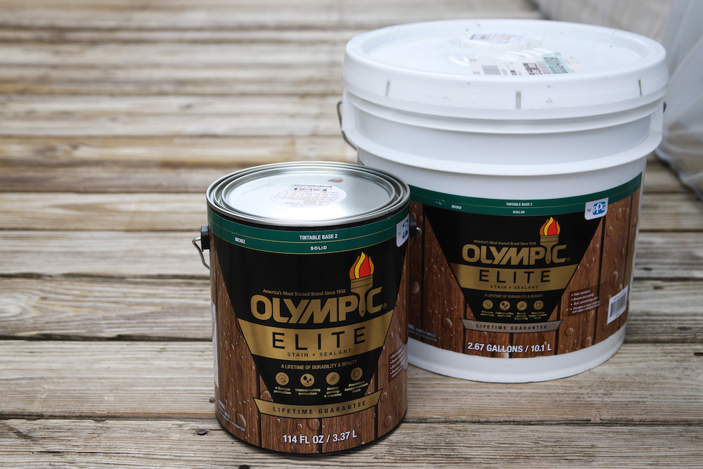 A can and a bucket of Olympic elite solid stain sitting on a deck. A can and a bucket of Olympic elite solid stain sitting on a deck.