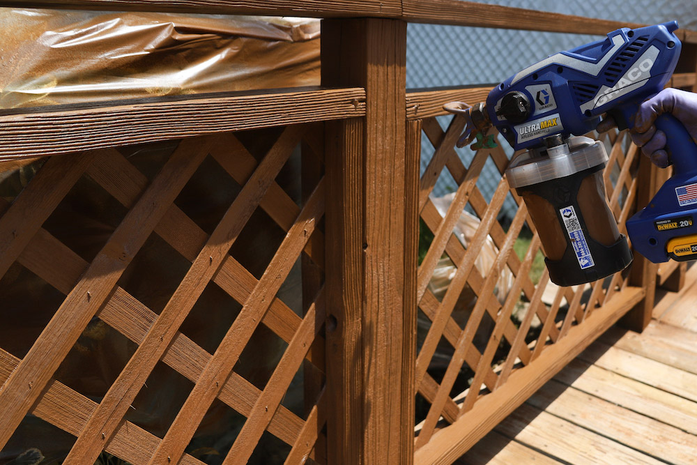 A person uses a paint sprayer to stain lattice and railings on a deck. A person uses a paint sprayer to stain lattice and railings on a deck.