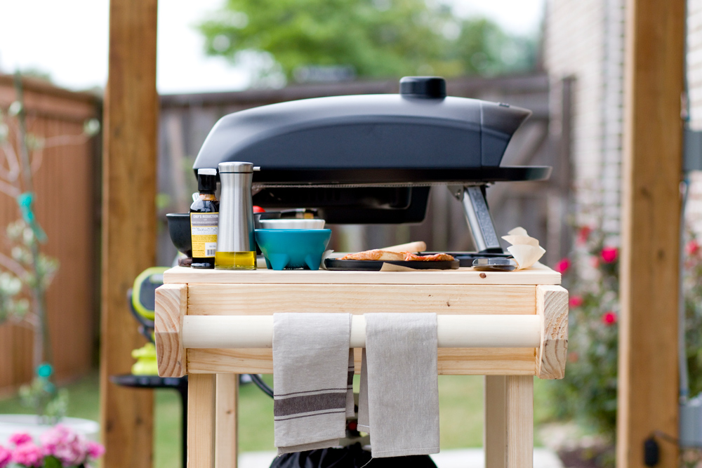 A pizza oven behind an assortment of pizza ingredients, on top of a pizza cart.