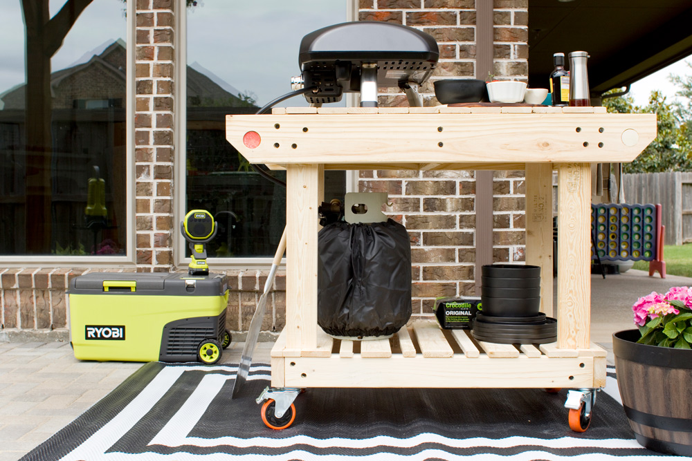 A rolling cart with pizza oven and ingredients on top, and a Ryobi kit behind. 