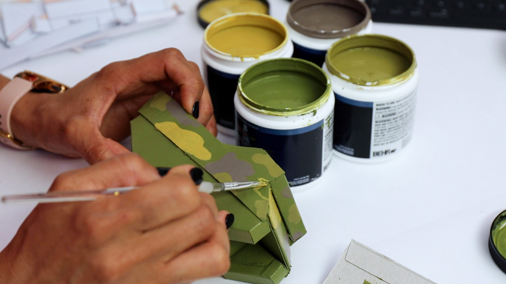 Hands painting a camo pattern on a DIY cardboard vehicle with containers of green paint on a table
