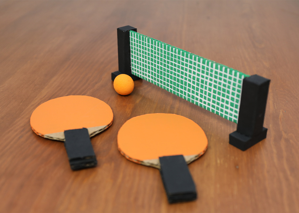 Two orange paddles, one small orange ball and a DIY tennis net placed on a wooden table top 