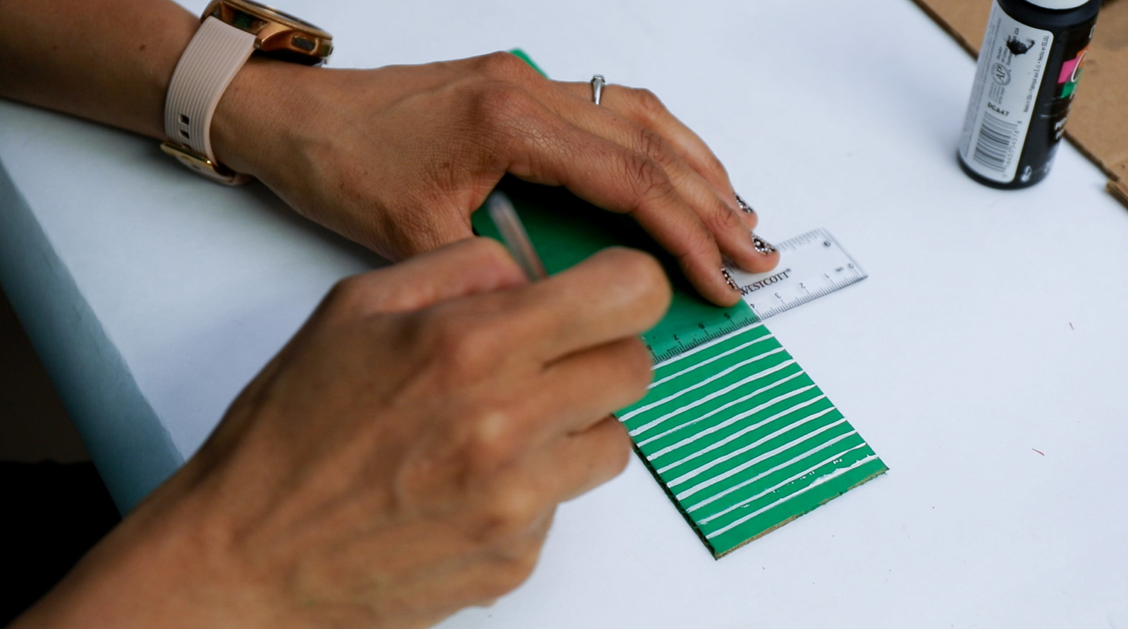 A person using a ruler to draw white lines on a green painted piece of cardboard 