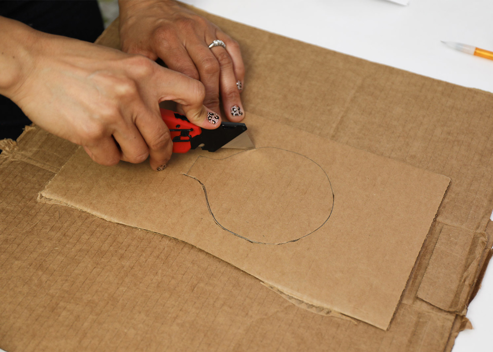 A person cutting out a traced outline on a piece of cardboard with a box cutter