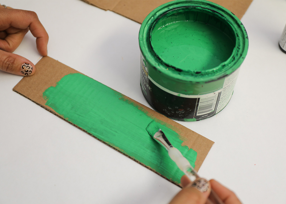 A person painting a piece of cardboard green with a small can of green paint in front of them 