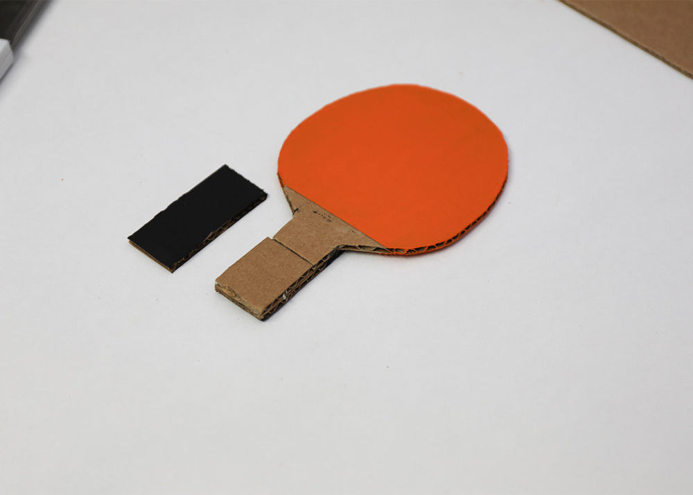 A DIY orange paddle laid on a white table top next a small black painted piece of cardboard