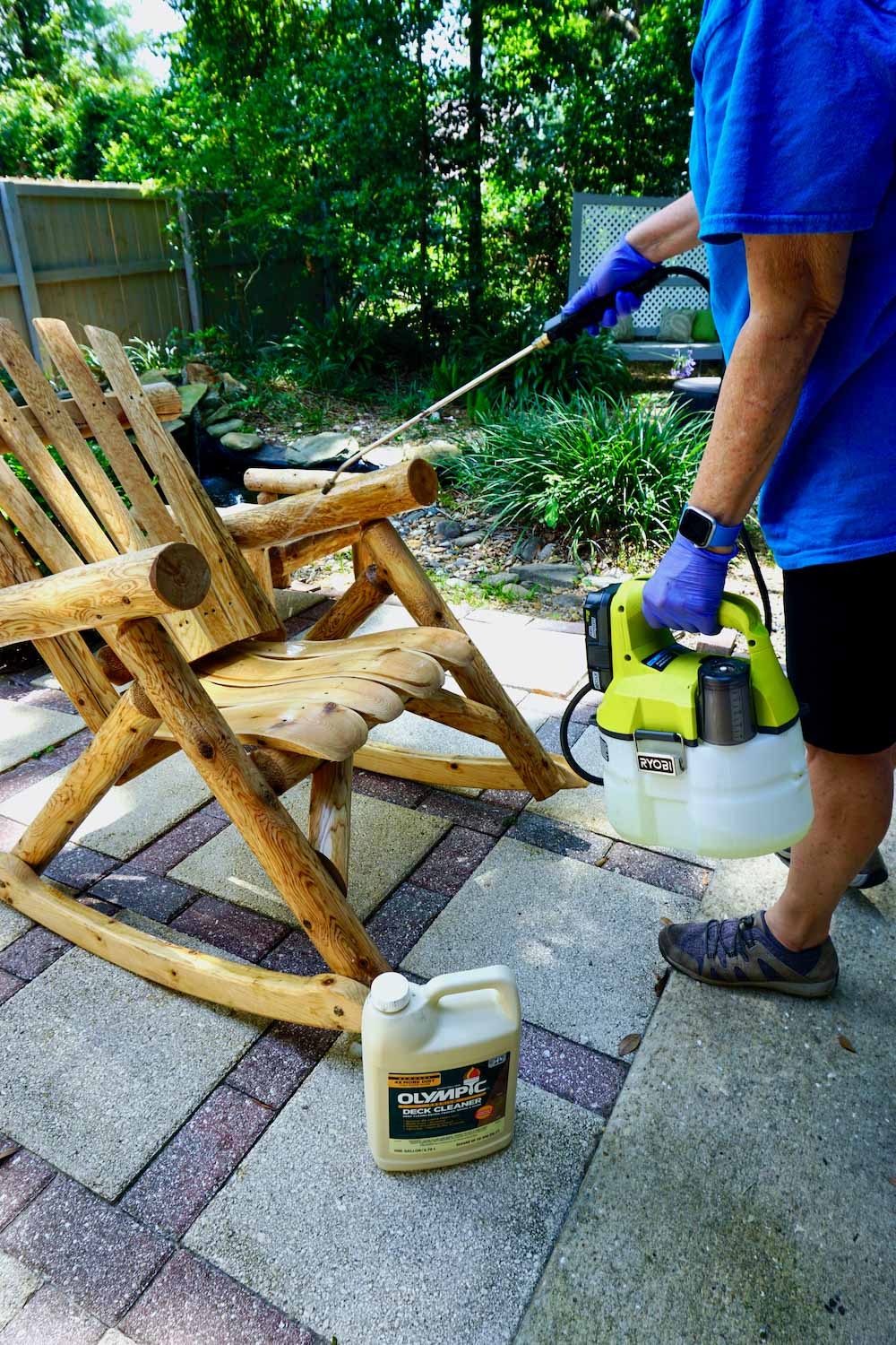 A person uses a pump spray filled with Olympic Premium Deck Cleaner to clean a wooden chair. A person uses a pump spray filled with Olympic Premium Deck Cleaner to clean a wooden chair.