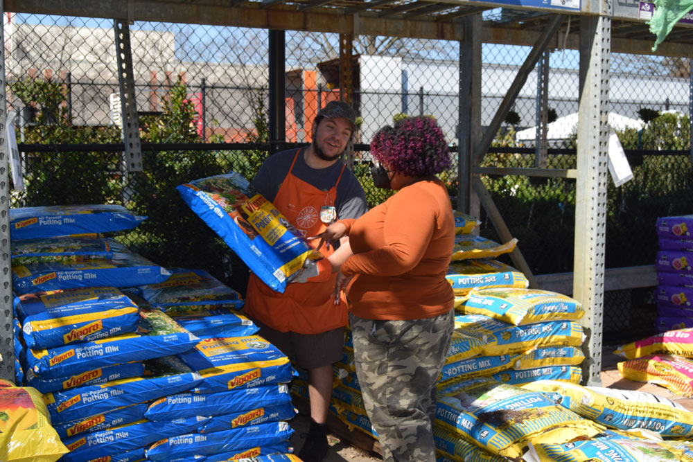 A woman speaking to a Home Depot employee holding a bag of Vigoro soil.