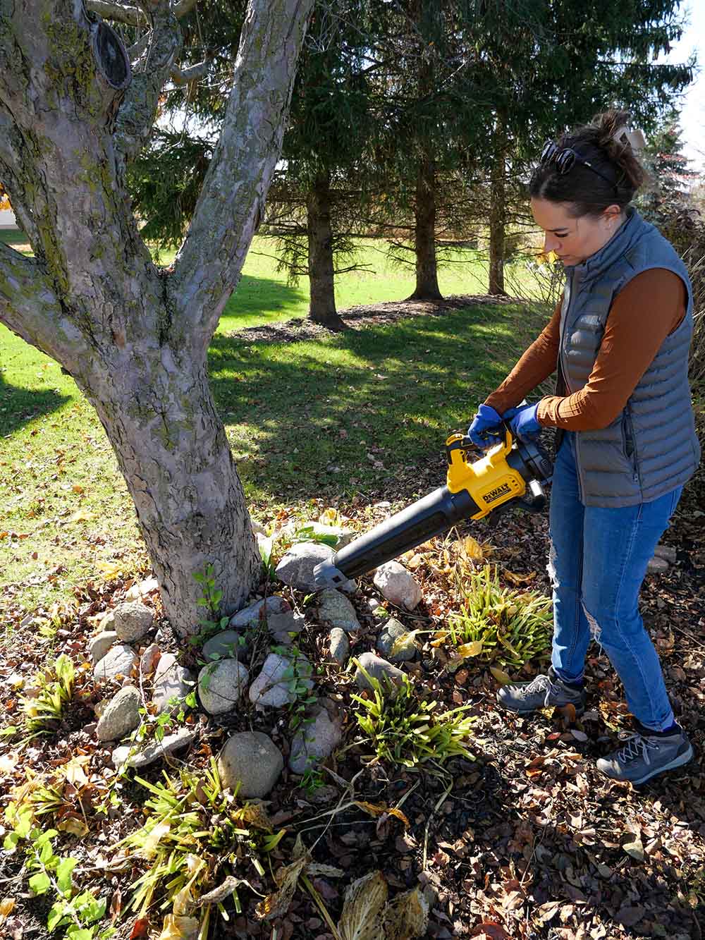 A woman using a leaf blower to remove scattered leaves from the garden beds.