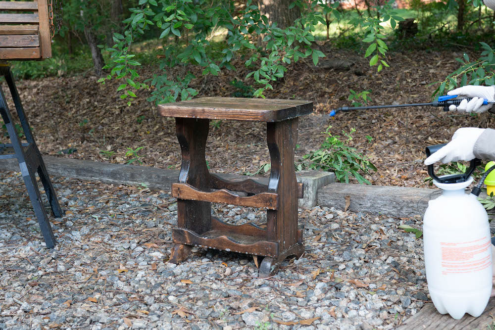 A pump sprayer applies Olympic Premium Deck Cleaner to a wooden side table.