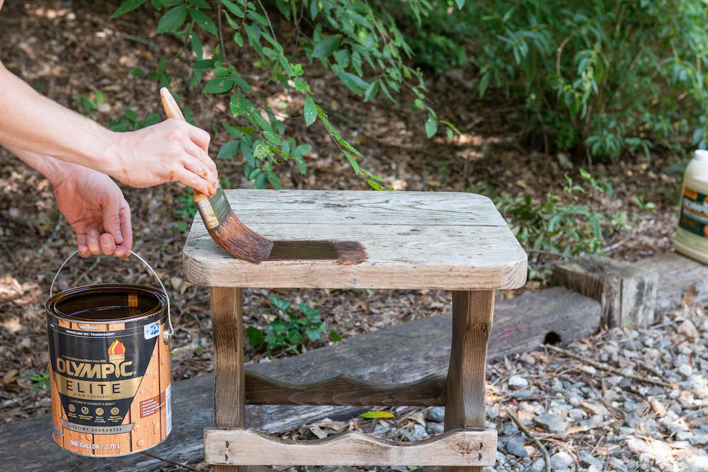 A person applies a coat of Olympic Elite Stain and Sealant to a wooden side table.