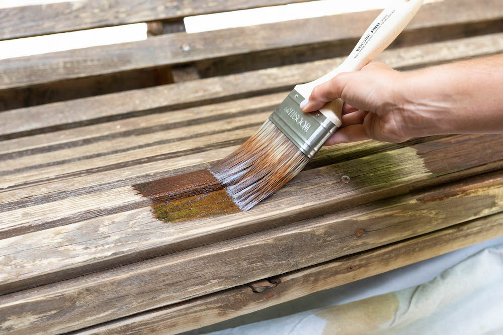 A person uses a paint brush to apply Olympic Elite Stain and Sealant to an exterior swing.