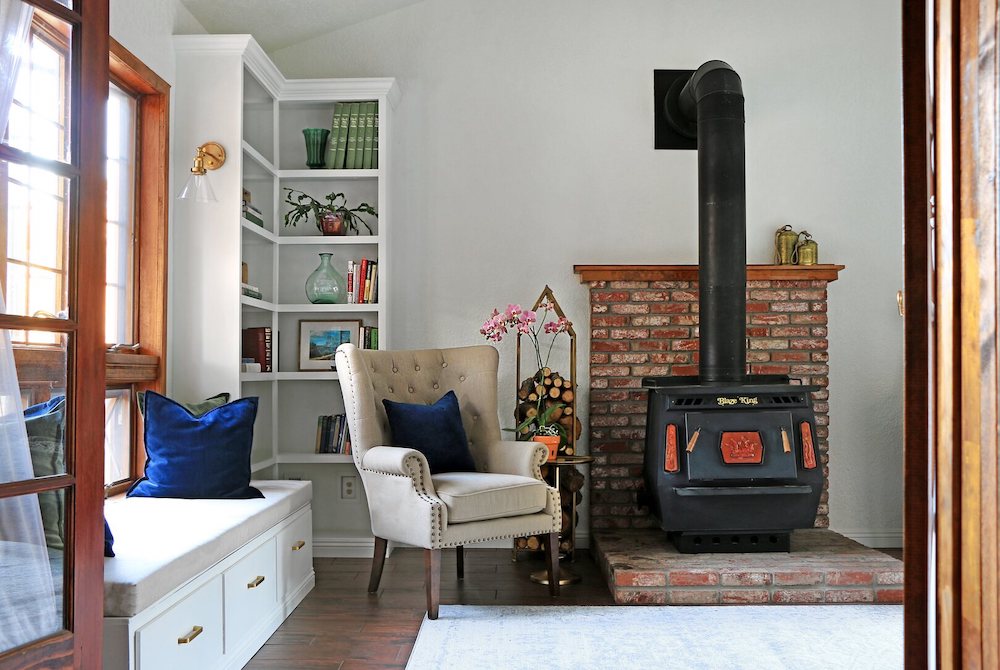 Beige armchair with a blue pillows placed in front of a corner bookshelf with an antique fireplace to the right and window bench with blue and green pillows to the left