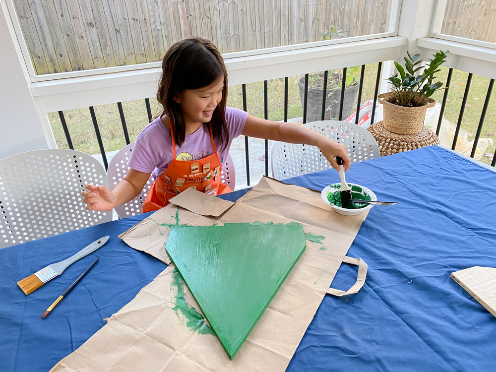 A girl using green paint to paint a wooden piece.