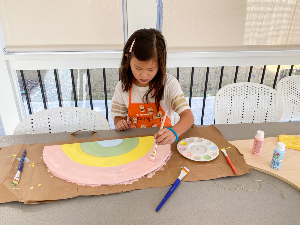 A girl using pink pastel paint to paint a rainbow on a piece of plywood.