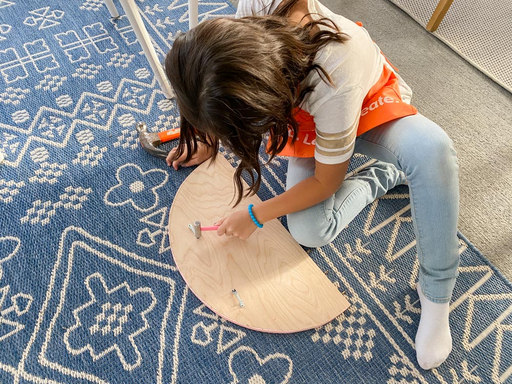 A girl sitting on the ground using a small hammer to nail hooks onto the back of a piece of plywood.