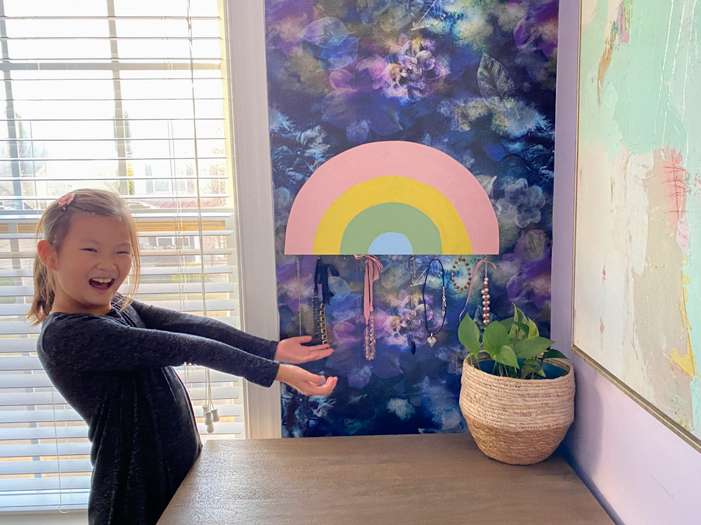 A girl presenting a DIY Rainbow Jewelry Holder project hanging on a wall.