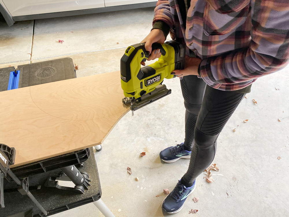 A woman using a jigsaw to cut a piece of plywood.