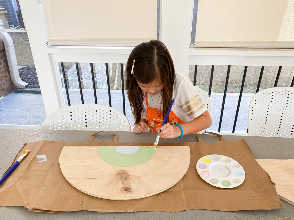 A girl using green pastel paint to paint a piece of plywood.