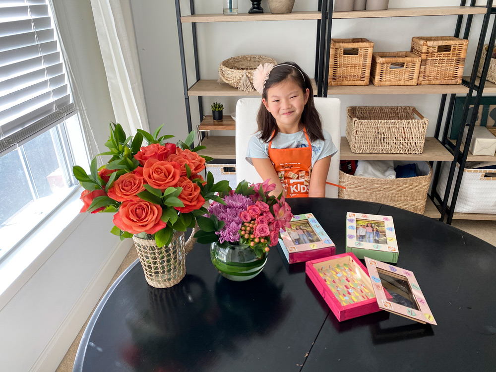 A girl sitting in front of a table with flower bouquets and four finished and painted Valentine’s Day Boxes.