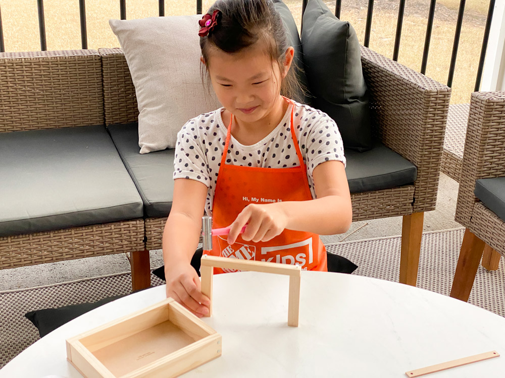 A girl nailing the top of a wooden frame for a Valentine’s Day box.