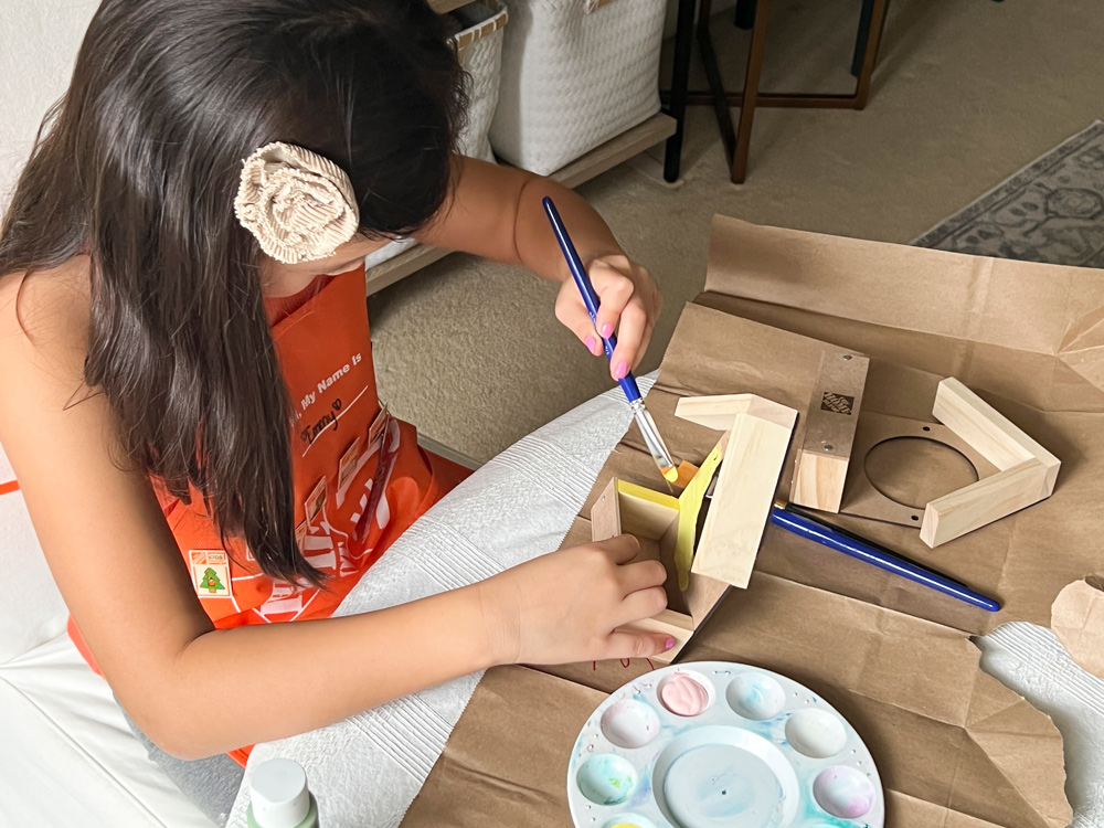 A girl using a paintbrush with yellow paint to paint a window bird feeder.