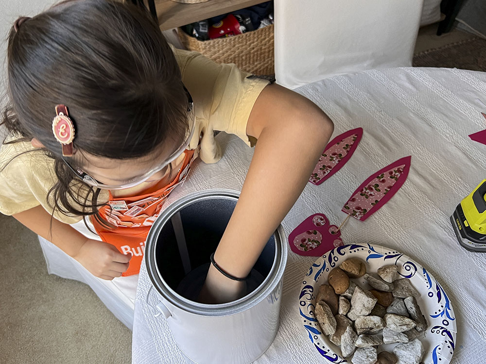 Girl reaching her hand in paint bucket with plate of rocks on table. 