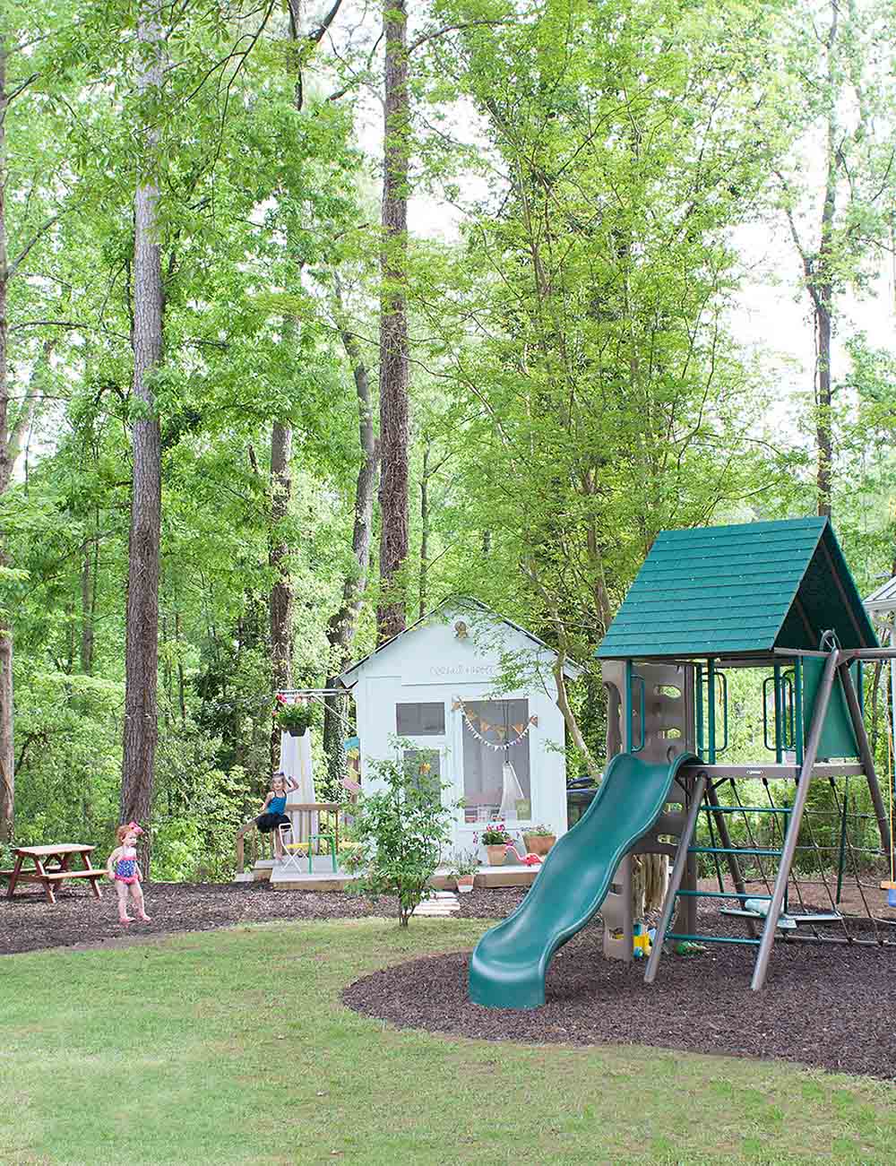 Two young girls play in a backyard next to a swing set and playhouse.