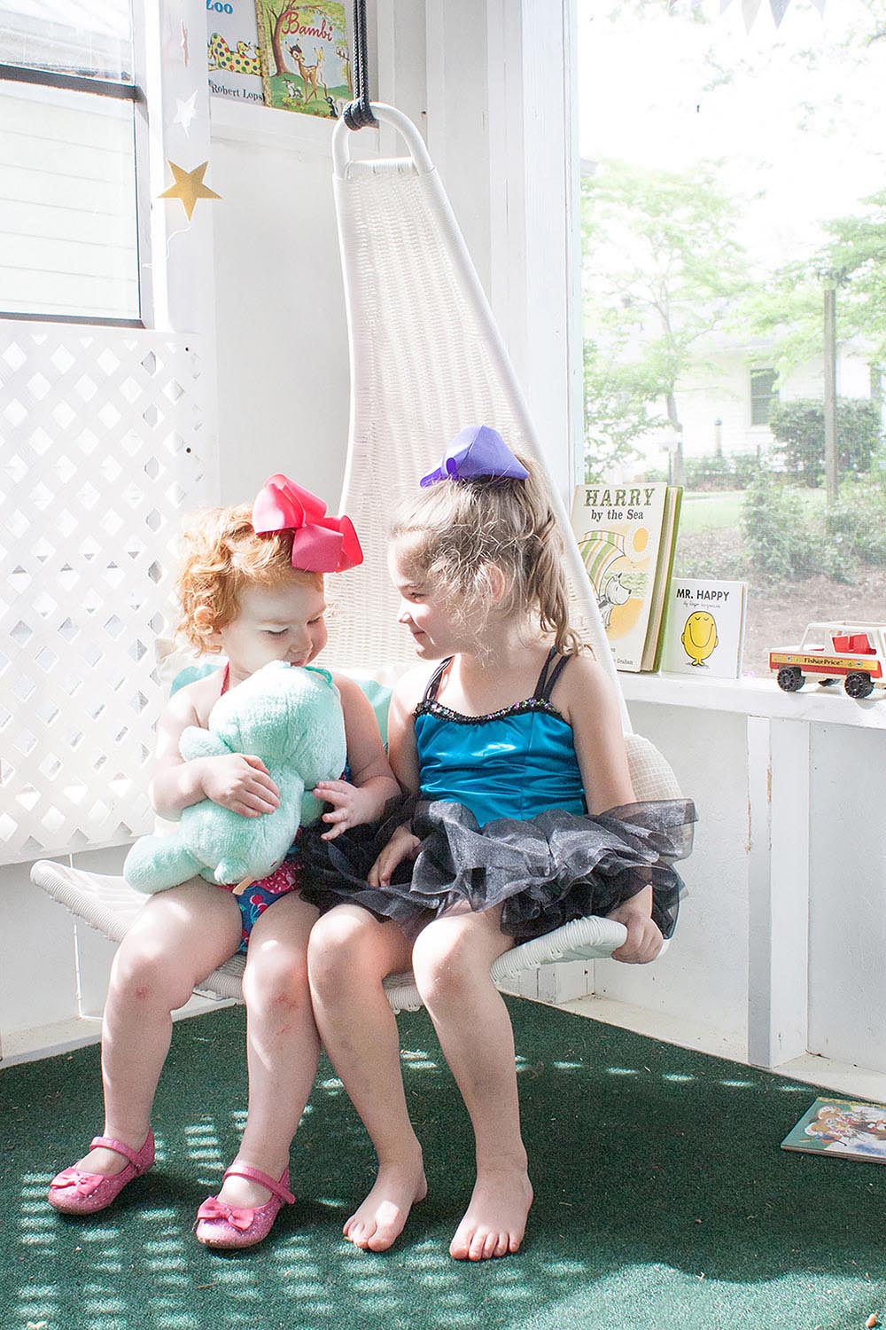 Two young girls sit in a white chair next to a window inside a playhouse.