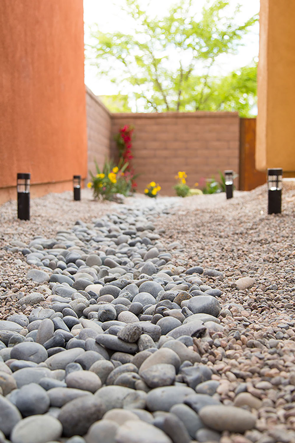 A completed DIY yard drainage made from Mexican beach pebbles and pea gravel is decorated with landscape lighting and plants.