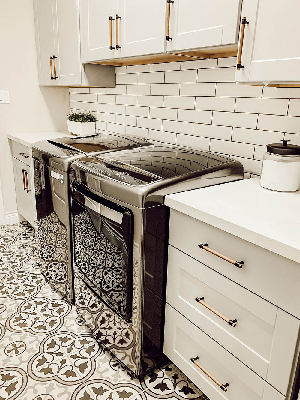 A laundry with grey cabinets and gold hardware, mosaic titled flooring, and a set of washer and dryers.