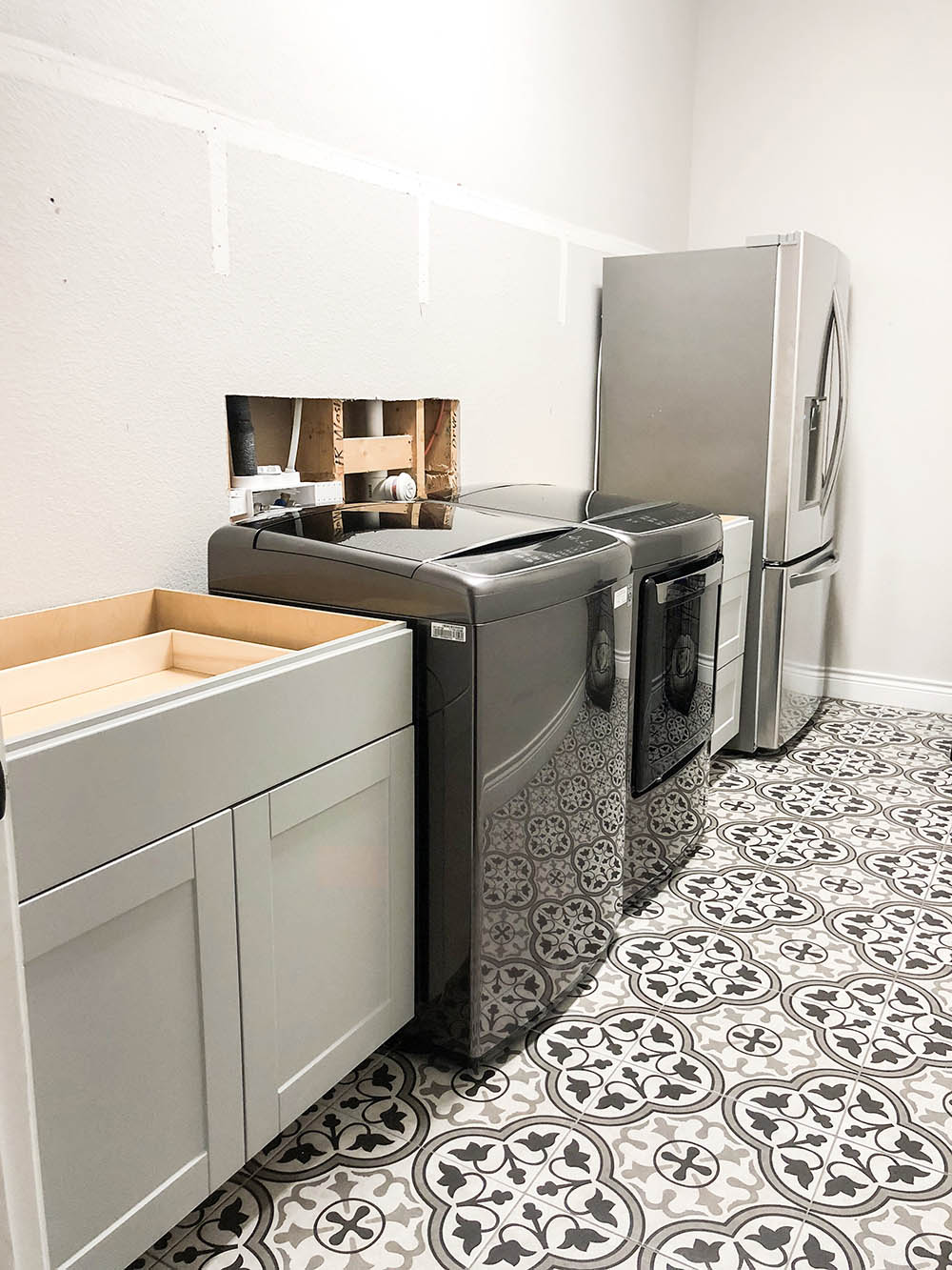 Laundry room with mosaic tiled flooring, black washer and dryer, grey cabinet and a refrigerator. 