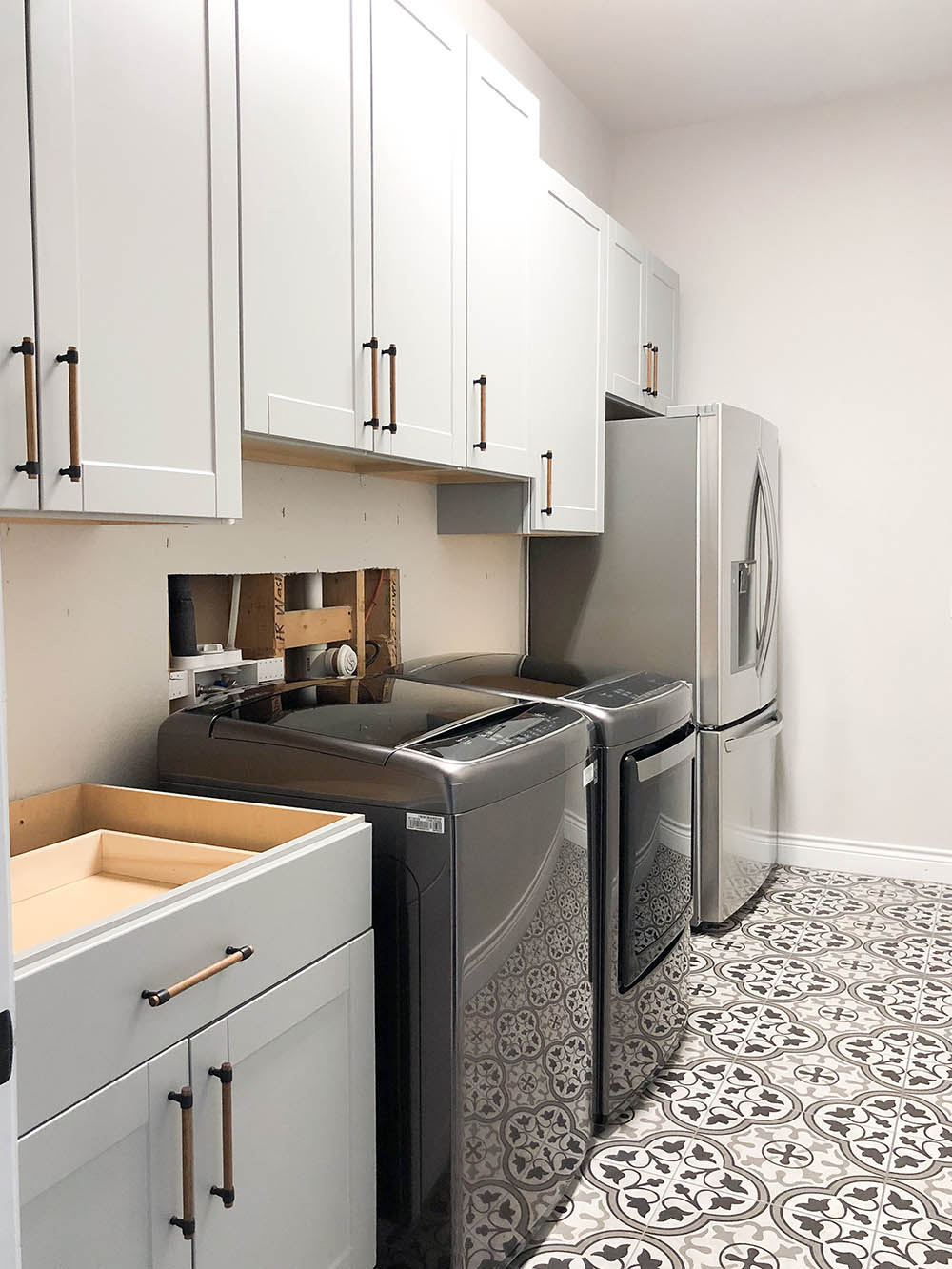 Laundry room with mosaic tiled flooring, black washer and dryer, upper and lower grey cabinets,  and a refrigerator. 
