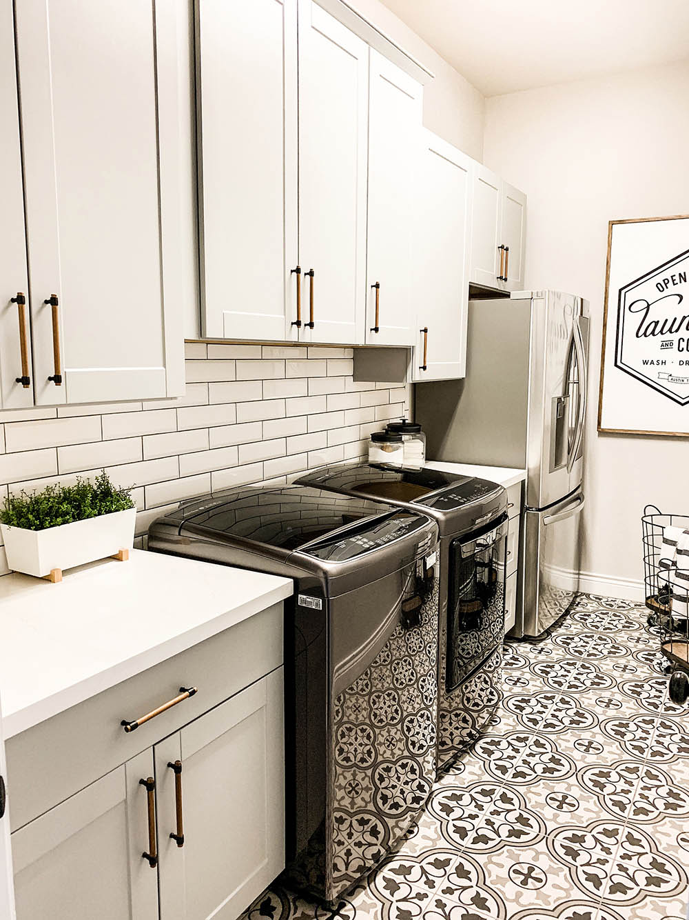 Laundry room with mosaic tiled flooring, black washer and dryer, upper and lower grey cabinets with gold hardware, subway tiled wall, wall art, and a refrigerator. 