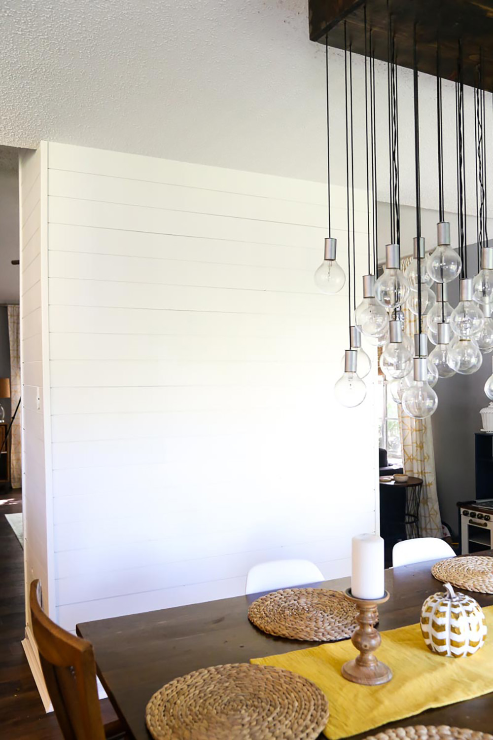 A white shiplap wall in a dining room with a wooden dining room table, placemats, candles and a handing light fixture.