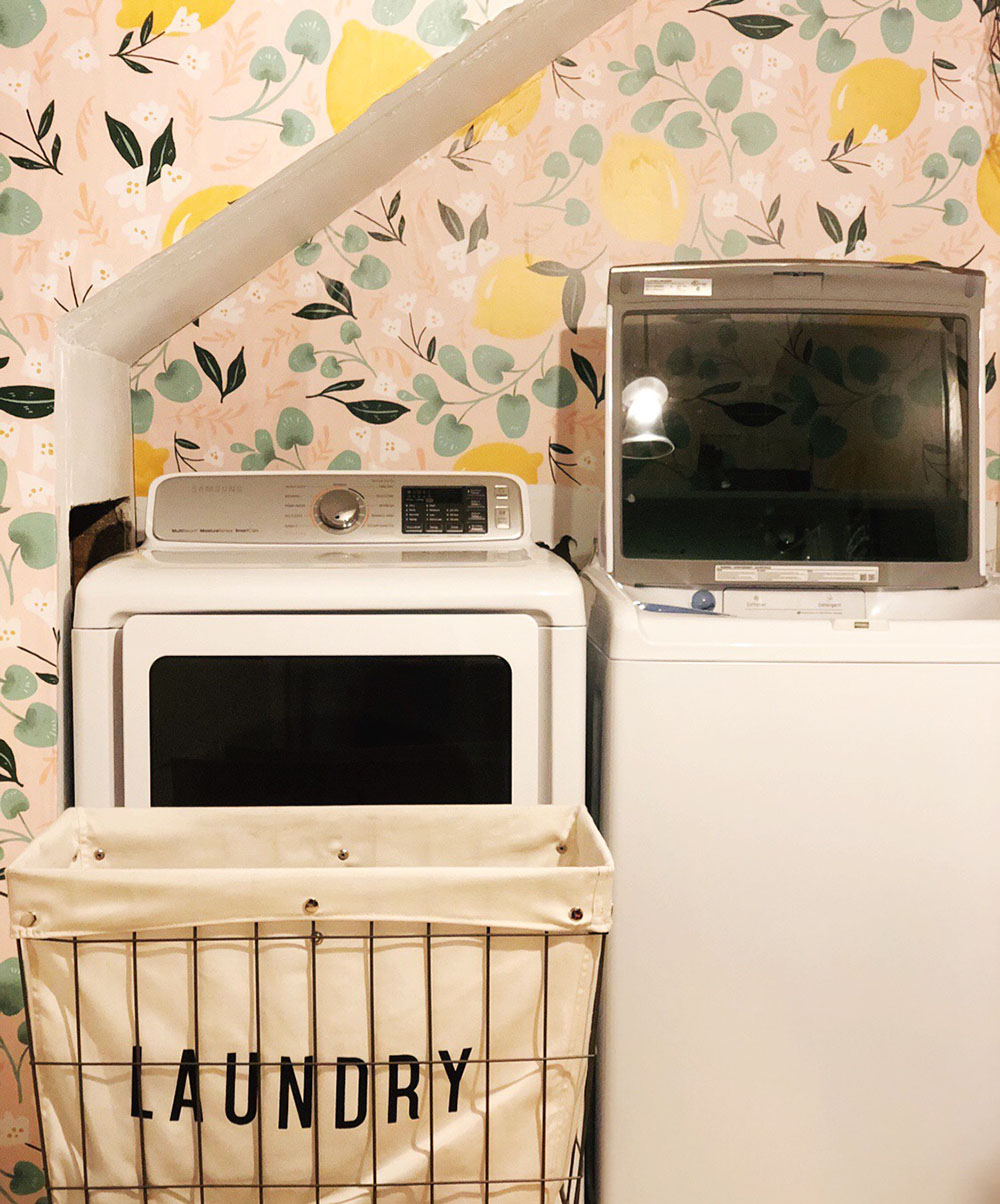 A wire and canvas laundry cart sits in front of a white washer and dryer set.