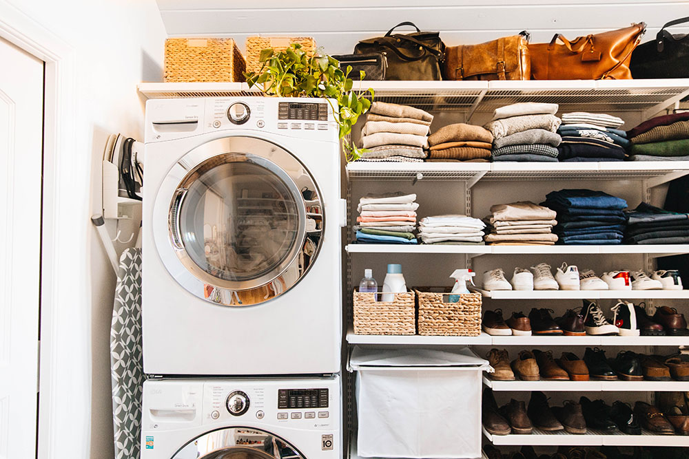 A small laundry room with shelves of clothing and shoes next to a stacked washing machine and dryer.