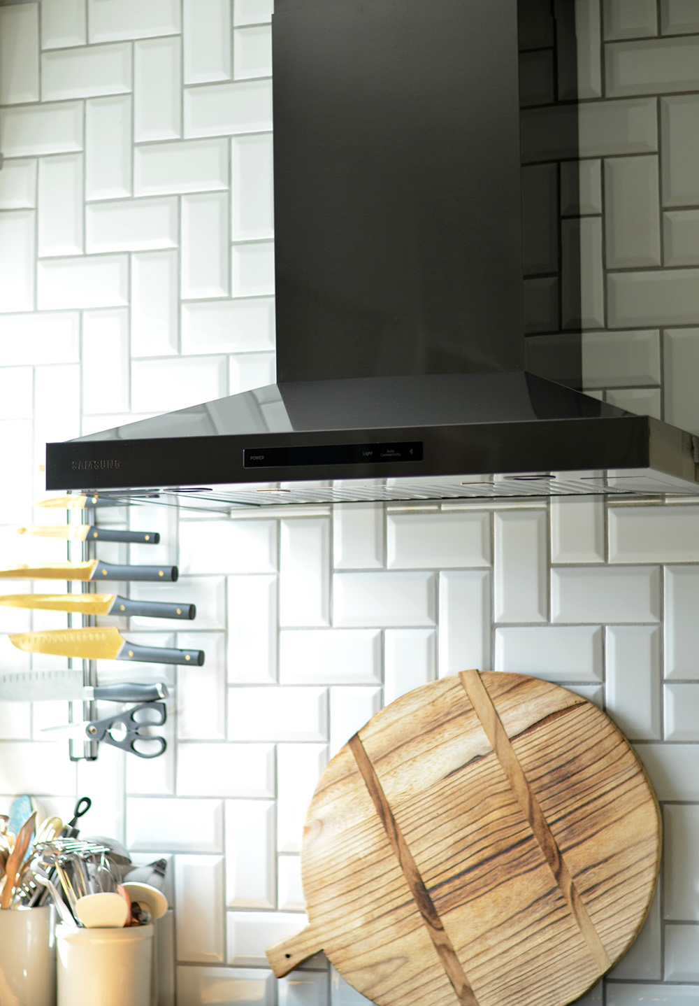 A black wall mount range hood hanging in front of a white tile backsplash.