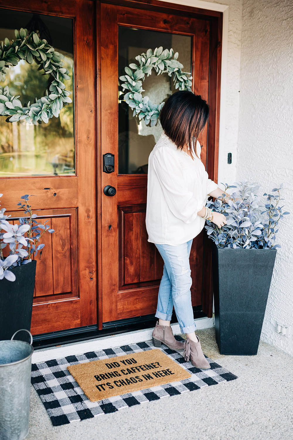 A woman standing in front of a renovated entryway fixes the greenery in a black planter.