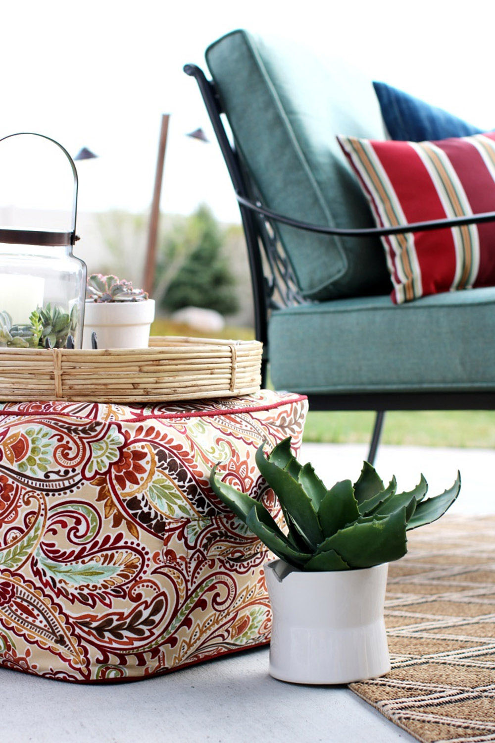 A wooden tray decorated with plants sitting on a paisley outdoor pouf. A wooden tray decorated with plants sitting on a paisley outdoor pouf.