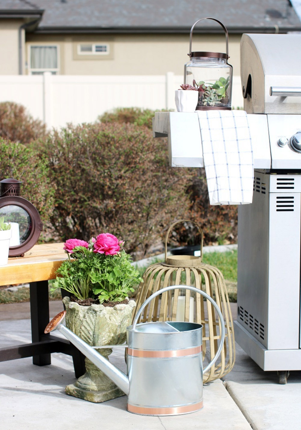A silver metal water can with copper accents sitting on the ground next to a grill. A silver metal water can with copper accents sitting on the ground next to a grill.