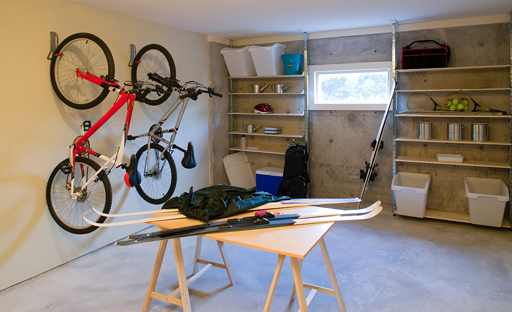 In a basement, bikes hang on hooks, sports equipment lays on a table and shelves line the back wall. 