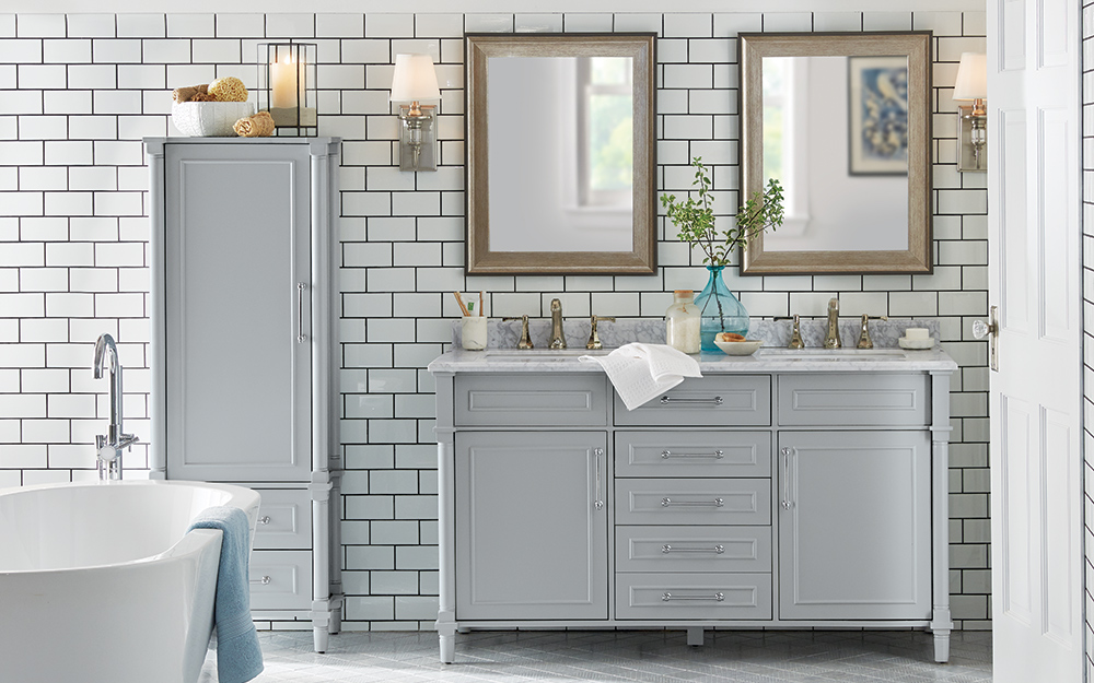 A bathroom features a freestanding tub, double vanity with matching gray upright storage cabinet and a tiled wall