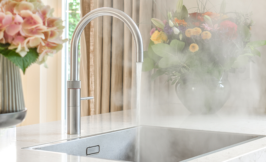 Steam rises as hot water runs from a faucet into a sink next to fresh flower arrangements.